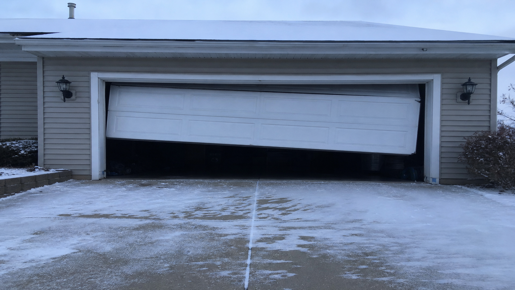 A residential garage door sitting slightly crooked and partly open over a snow-dusted driveway apron on a cold Illinois morning