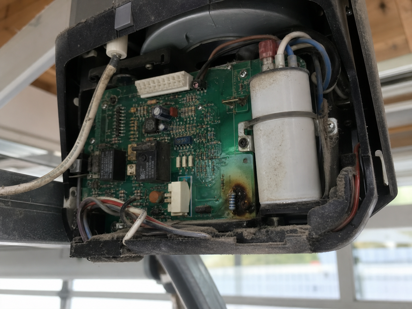 Close-up of an opened garage door opener motor head showing a green circuit logic board with a discolored, heat-scorched component near the capacitor