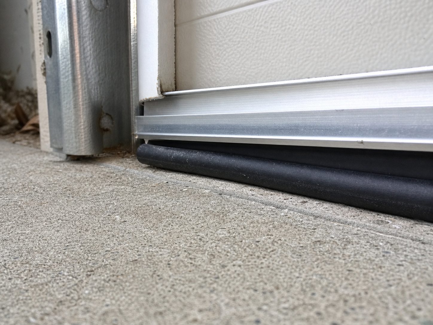 Close-up of the black rubber bottom weather seal on a garage door pressed against a concrete floor threshold, with the aluminum retainer above