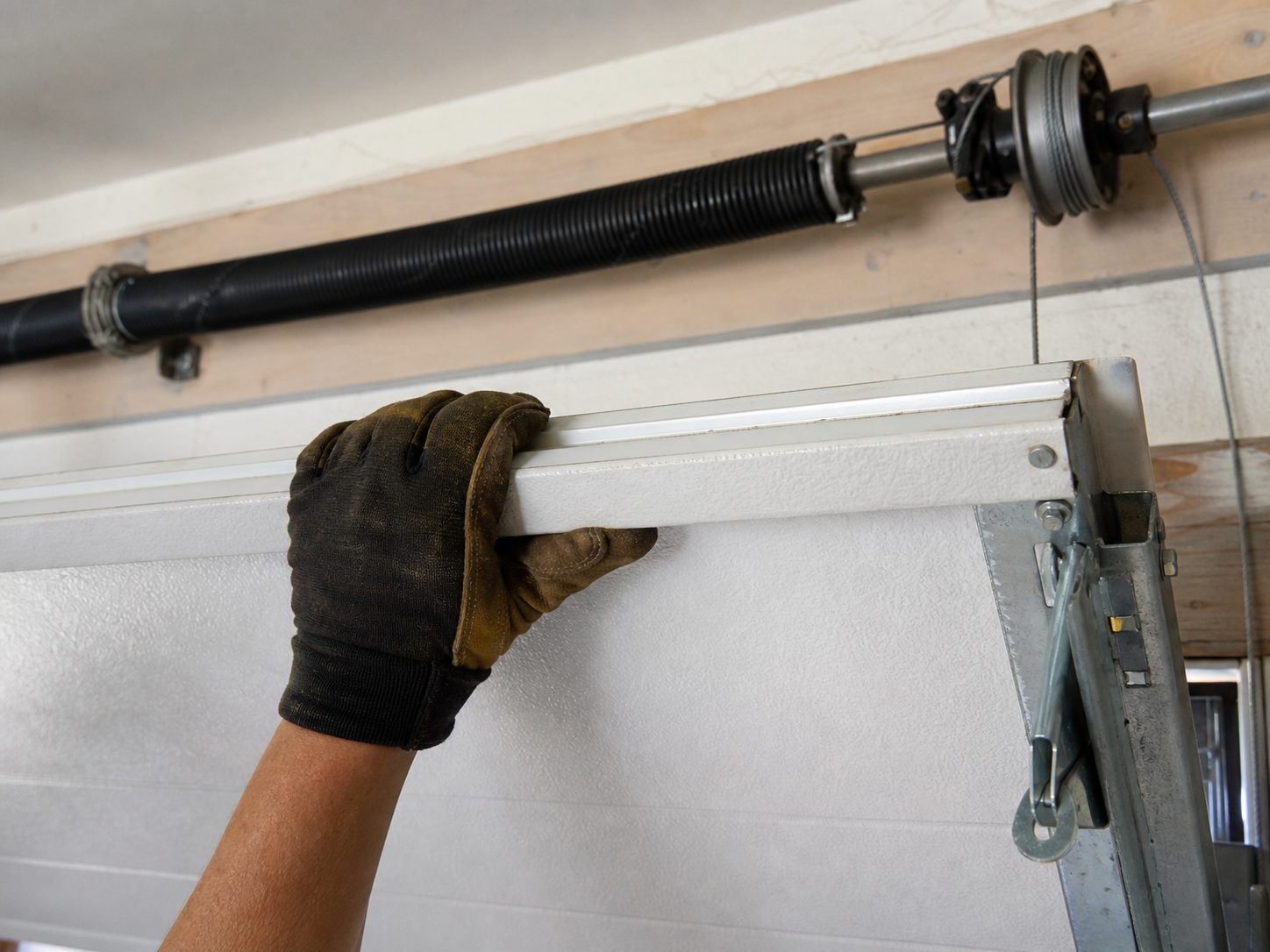 Technician's gloved hand steadying a partly open insulated garage door during a balance check, with the black torsion spring and cable drum on the shaft above